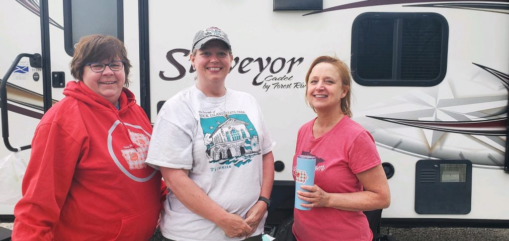 Three women standing in front of a camper, one wearing a red hoodie and the other holding a blue travel mug.