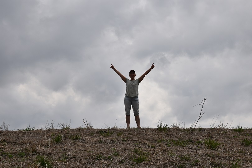 I woman at top of a hill on a cloudy day with her arms in the air celebrating her arrival at the top.