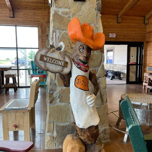 A six foot tall wooden carving of a sausage in an orange cowboy hat wearing an apron with the name Big H.