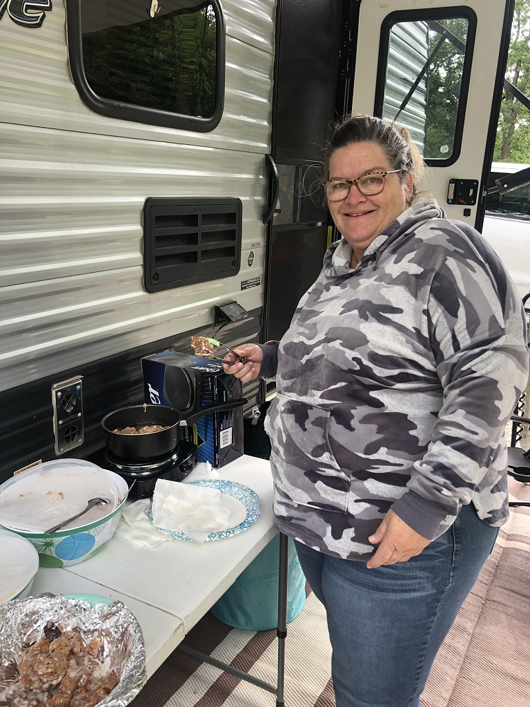 A woman wearing a camo hoodie making apple fritters on a table outside her camper.