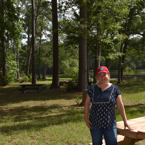 Lisa Dempsey Girl Camper Southeast Texas Chapter Guide is standing next to a picnic bench at the park in her red Girl Camper hat