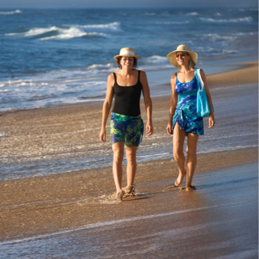 Two women in hats walk on galveston island beach.  One is carrying a blue bag and has dark sunglasses.