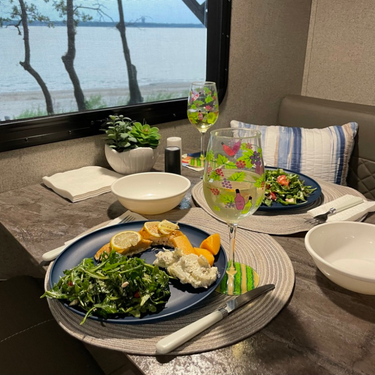 A dinner table set with wine glasses and a meal inside of a camper and a view of the lake outside of the window featuring cedar planked salmon
