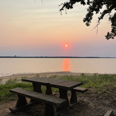 A campsite with a picnic table overlooking a lake at sunset.
