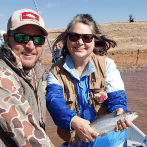 A man and a women are in a boat on a lake in Oklahoma.  The woman is holding a rainbow trout and a fishing pole. 