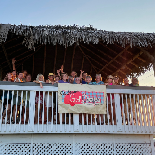 A large group of women are standing on a balcony under a thatched style roof holding a Welcome to Girl Camper Sign