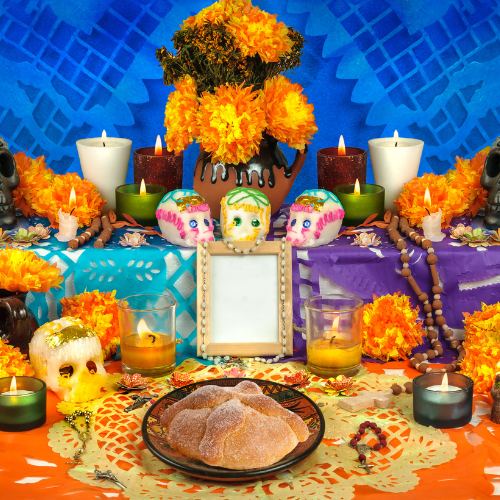 Pan de Muerto, a loaf of sugar sweet bread, is on a bright orange tablecloth surrounded by brightly painted sugar skulls and lighted candles to celebrate dia de los muertos or the day of the dead