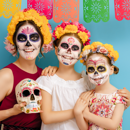 Three girls wearing flowered head bands with faces painted like colorful skulls holding a painted skull to celebrate dia de los muertos