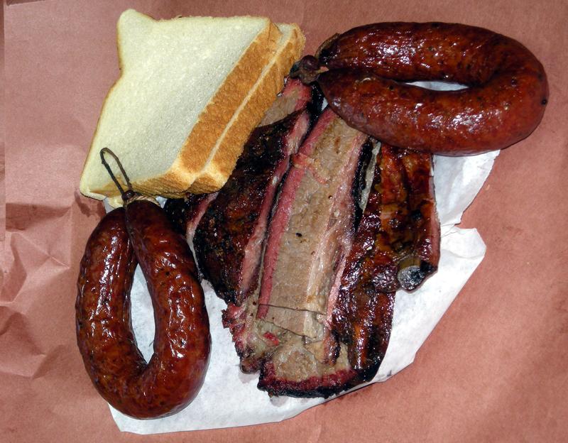 A sheet of pink butcher paper is filled with two slices of bread, sliced beef brisket, and two rings of link sausage from the City Market BBQ restaurant located near Palmetto State Park