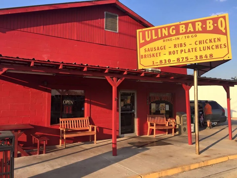 A Red building with a yellow sign reads Luling Bar-B-Q.  Two wooden benches sit on either side of the door