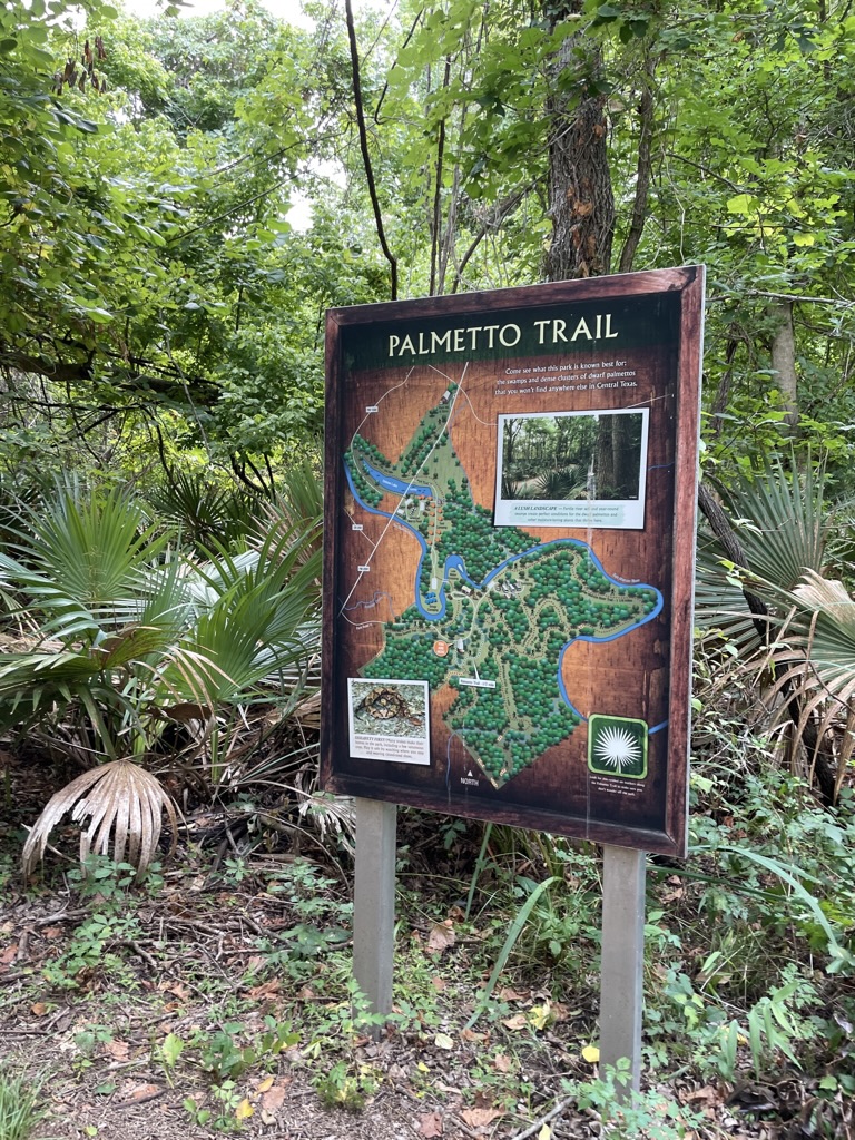 A sign notating Palmetto Trail and a map sits at a trailhead in the trees