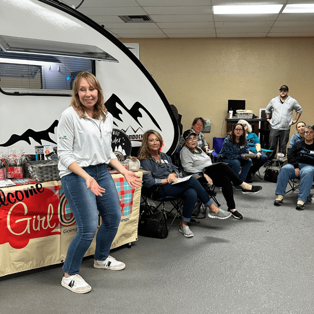 Sue Ann with Princess Craft RV welcomes a group of women sitting in camp style chairs in front of a small nuCamp TAB trailer