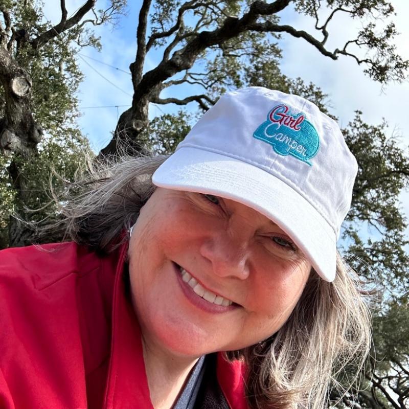 Lisa Dempsey is wearing a white baseball cap with girl camper logo and looking down at the camera.  Behind her is a large oak tree.