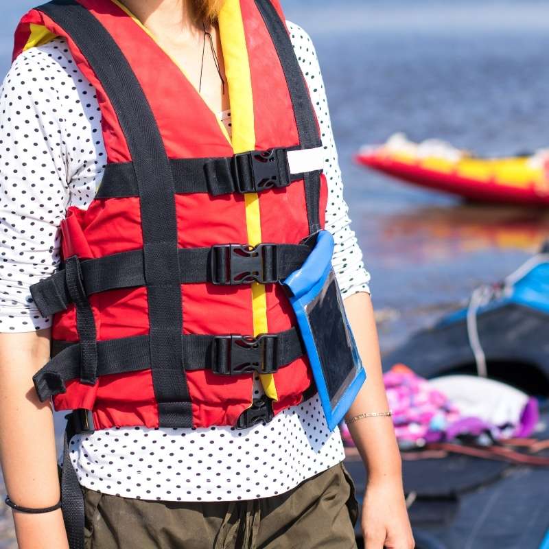 a women wearing a red life jacket stands by her kayak on the side of a river.