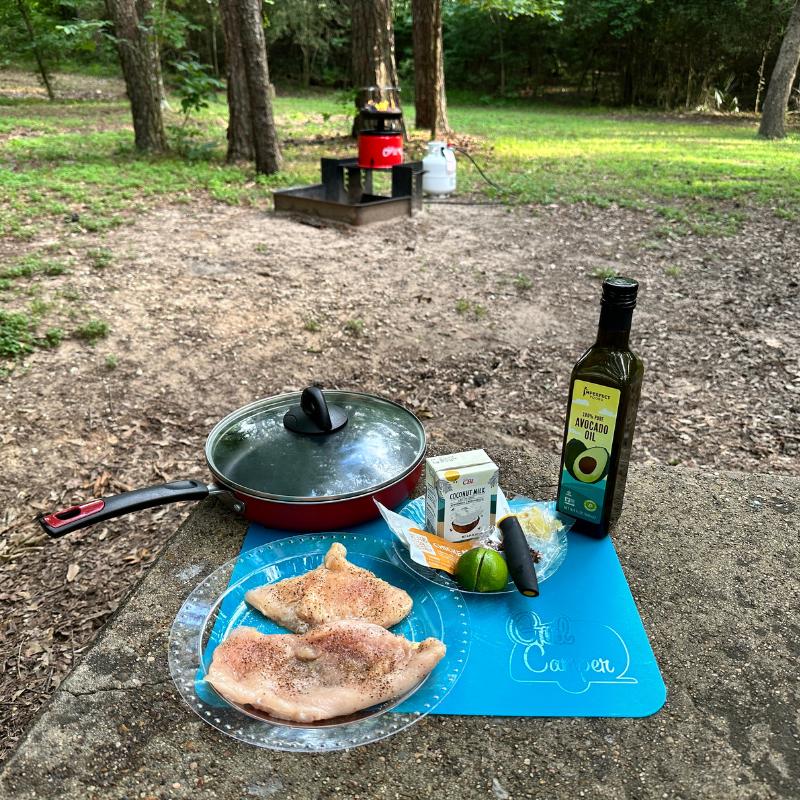 Camco Big Red Campfire in the background and a meal preparation of chicken, oil, a frying pan sit on a picnic table.