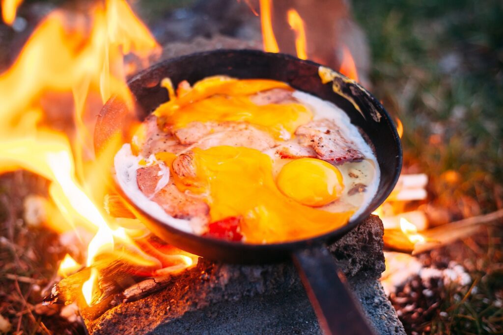 A picture of a cast iron pan filled with eggs and bacon on a campfire. 